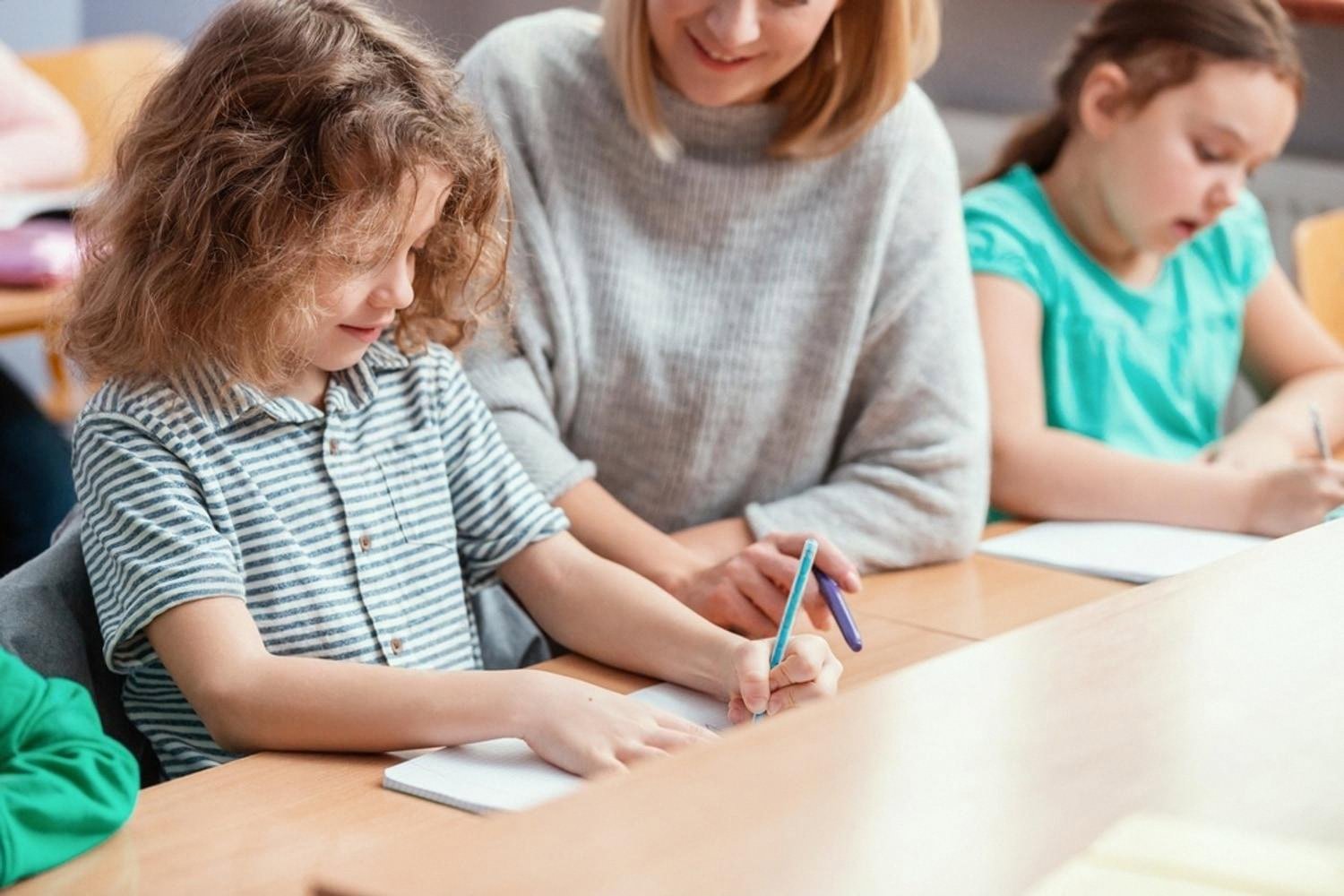 First-grader writing in a notebook while a teacher gently guides her hand in class