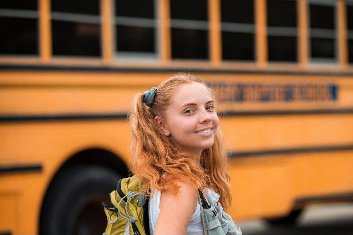 Smiling first-grader with a backpack standing next to a yellow school bus on the first day of school