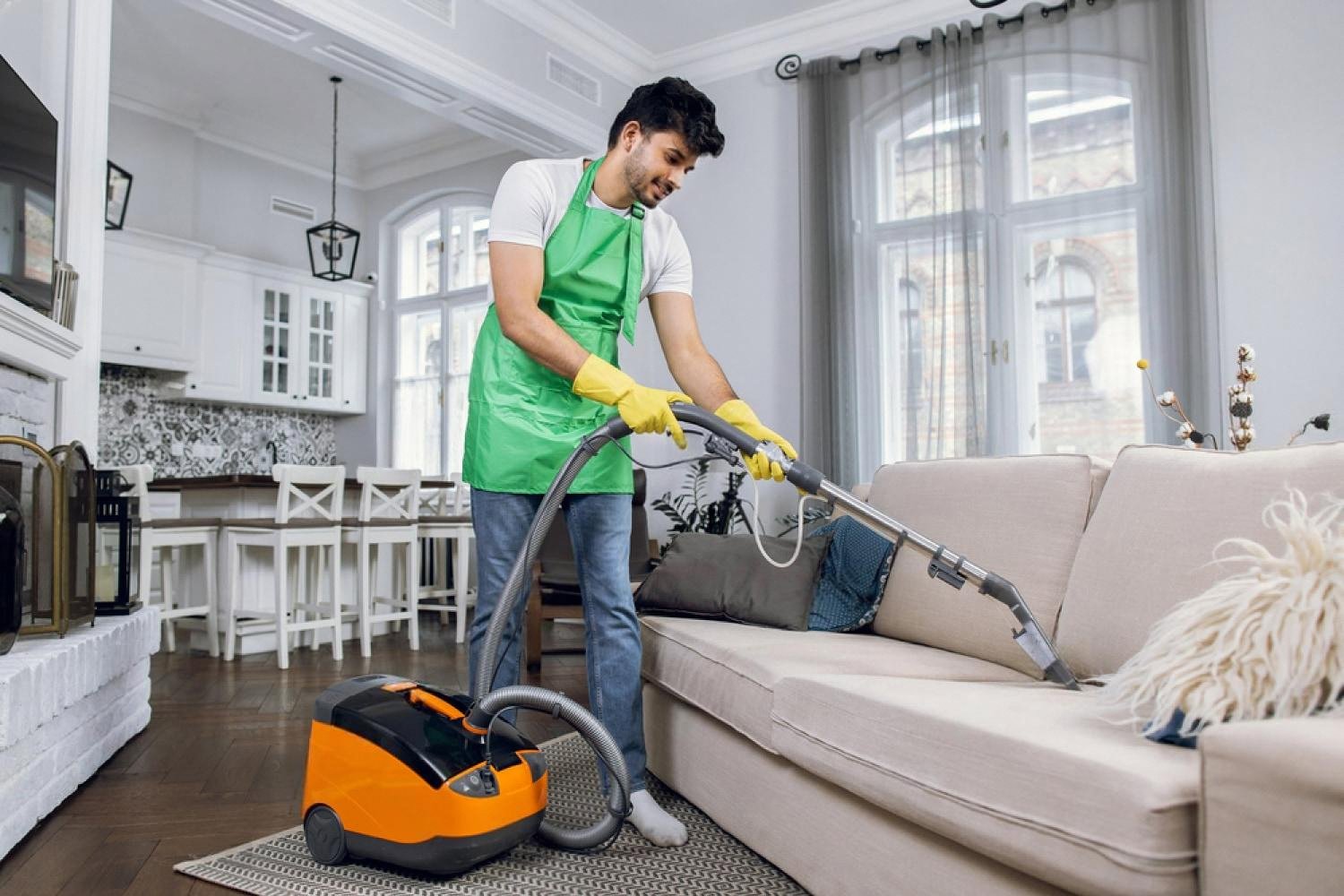 Professional cleaner in a green apron using an extraction vacuum on a sofa and rug in a Dubai home